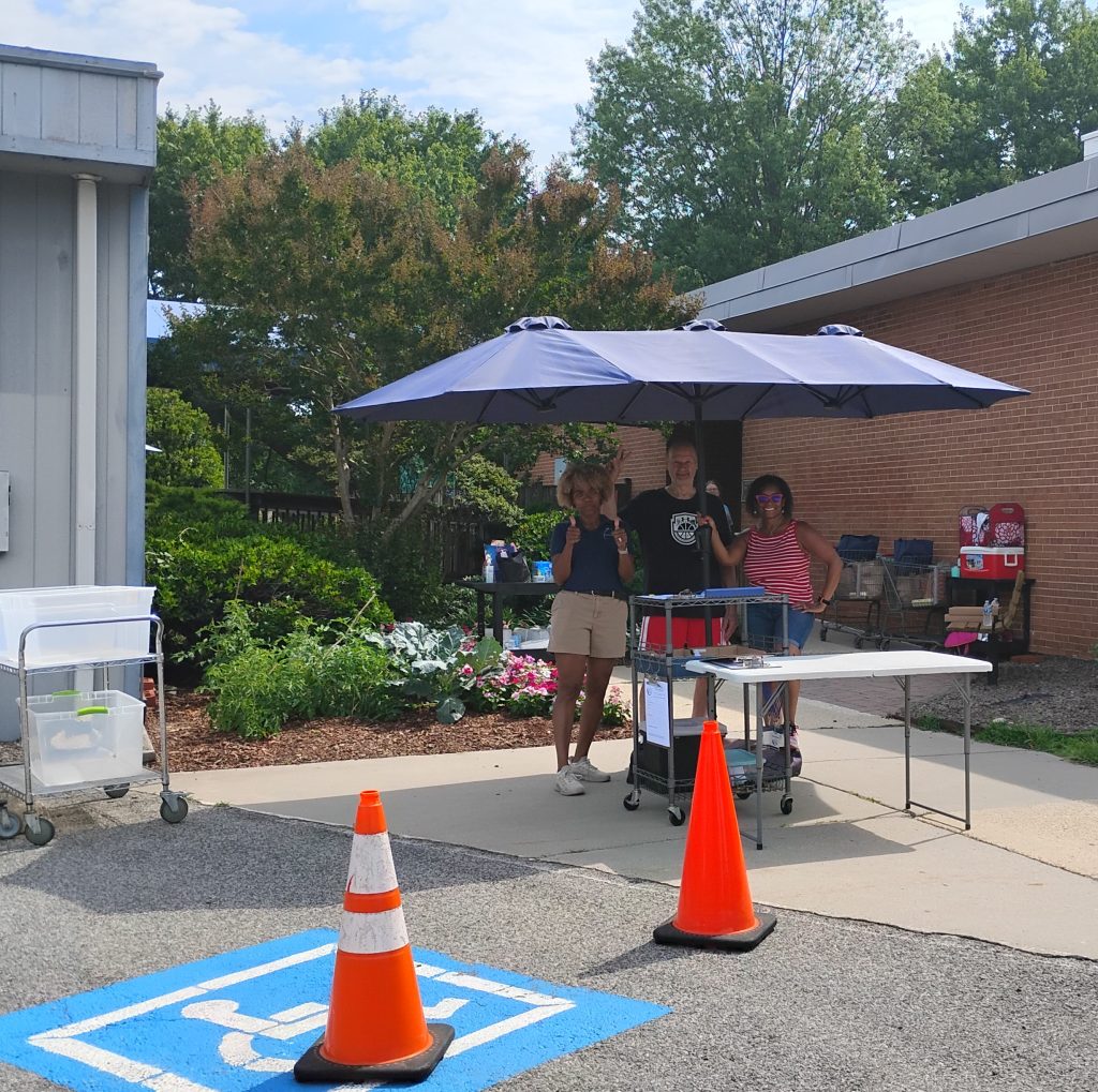 Three people stand under a large umbrella at an outdoor table with drinks, next to orange cones marking a handicapped parking spot. Green bushes and brick buildings are in the background.