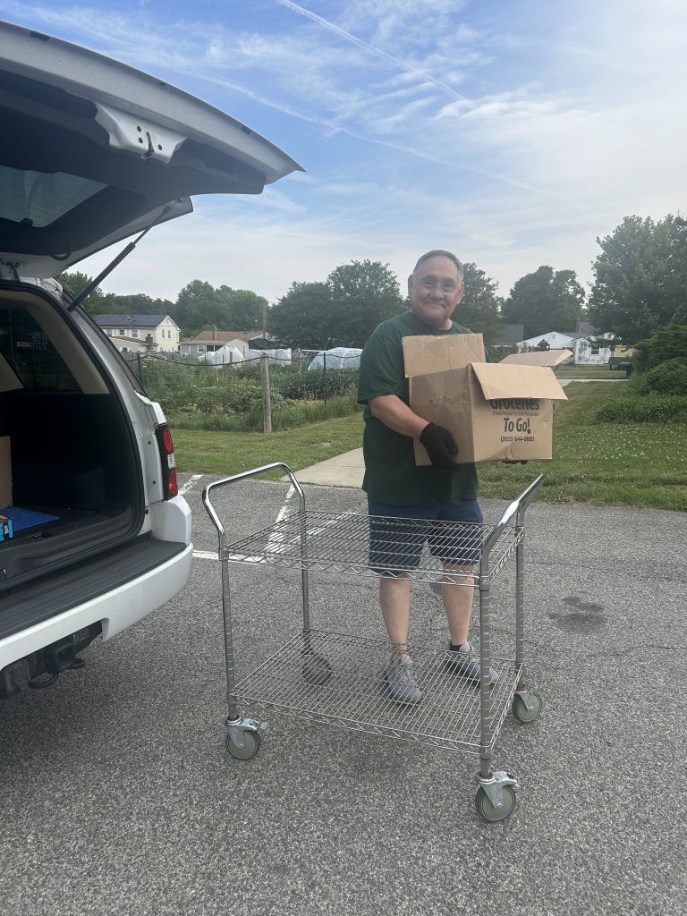 A person stands outside by a metal cart, holding two cardboard boxes and smiling. An SUV with an open trunk is parked nearby, and houses and trees are visible in the background under a partly cloudy sky.