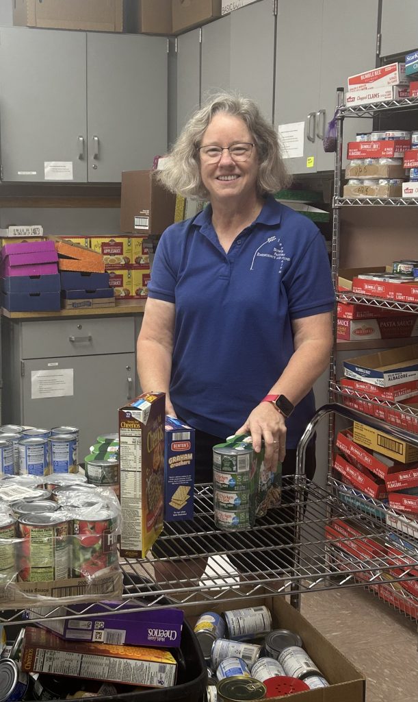 A woman with gray hair and glasses, wearing a blue polo shirt, smiles while organizing canned goods and boxed food on a metal cart in a storage room filled with shelves of food.