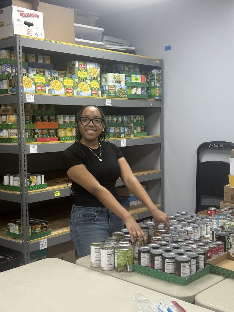 A woman smiles while organizing canned goods on a table in a food pantry. Shelves filled with canned foods are visible behind her.
