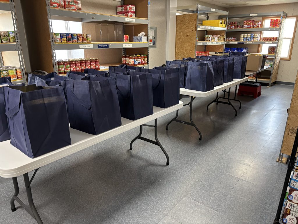 Reusable navy blue shopping bags filled with items are lined up on two white folding tables in a room with shelves stocked with canned and boxed food, suggesting a food pantry or donation center.